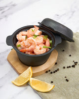 Black pot with shrimp on a marble surface with lemon wedges and peppercorns.