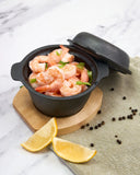 Black pot with shrimp on a marble surface with lemon wedges and peppercorns.