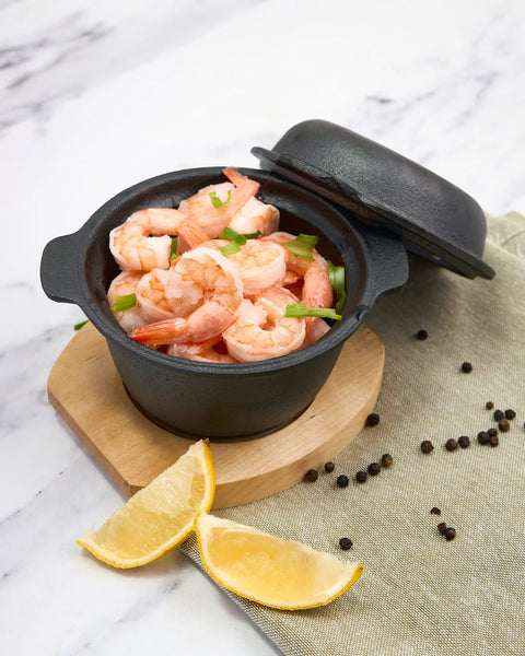 Black pot with shrimp on a marble surface with lemon wedges and peppercorns.