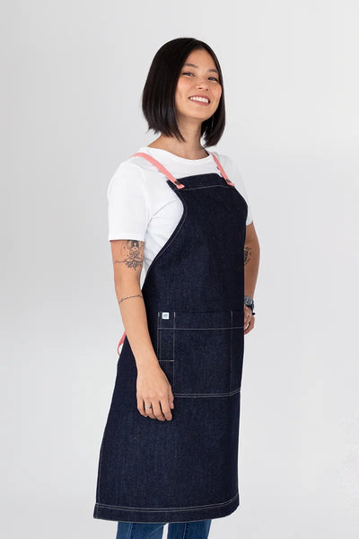 A person wearing a dark blue denim bib apron with grapefruit pink straps.