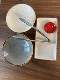 Two ceramic bowls and a small rectangle platter plate with a red ramekin and silver serving tongs. 