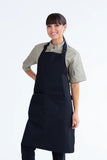 A person wearing a heavy weight black bib apron with an adjustable neck loop and waist ties, featuring a large front pocket with a pen insert.