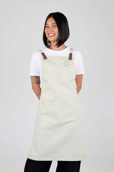 A person wearing a white bib apron with leather-look press stud straps and brass-coloured eyelets.
