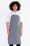 A person wearing a white shirt and a black bib apron with a striped pattern, featuring a pen pocket and an adjustable neck tie.