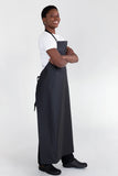 A person wearing a black bib apron with adjustable straps, standing with arms crossed.