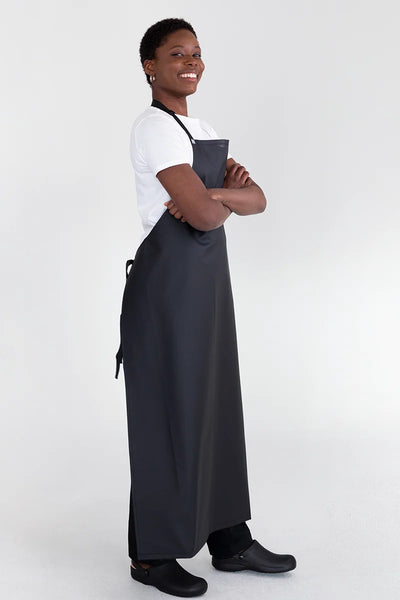 A person wearing a black bib apron with adjustable straps, standing with arms crossed.