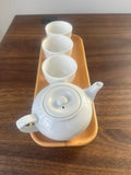 A white ceramic teapot with a lid, accompanied by three matching small cups, all arranged on a rectangular light wood tray over a dark brown wooden table.