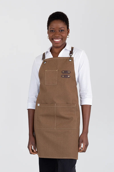 A person wearing a brown bib apron with leather-trimmed straps and utility pockets.